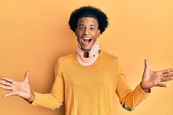 Afrikaanse amerikaanse man met afro haar dragen cervicale nek kraag vieren overwinning met vrolijke glimlach en winnaar uitdrukking met opgeheven handen  - Foto, afbeelding