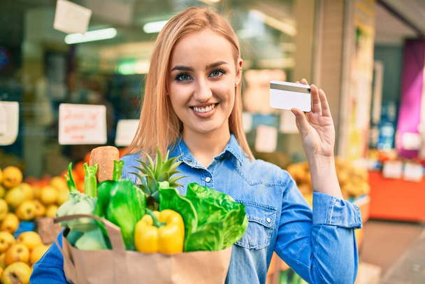 jong blond meisje glimlachen gelukkig vasthouden boodschappen papieren zak en credit card staan in de supermarkt - Foto, afbeelding