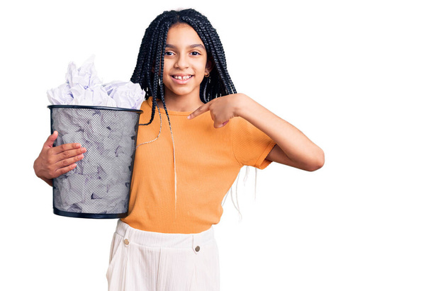 Симпатичная африканская американка с бумажной корзиной, полной скомканных бумаг, указывающая пальцем на одну улыбающуюся счастливую и гордую  - Фото, изображение