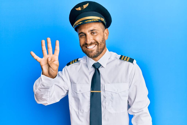 Hombre guapo con barba llevando uniforme de piloto de avión mostrando y apuntando hacia arriba con los dedos número cuatro mientras sonríe confiado y feliz.  - Foto, imagen