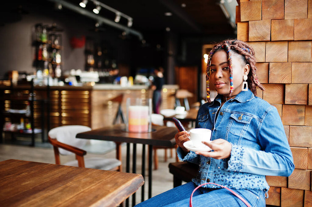 Lovely african american woman with dreadlocks in blue stylish jeans jacket at cafe hold mobile phone. Beautiful cool fashionable black young girl indoor drink coffee. - Foto, Imagem
