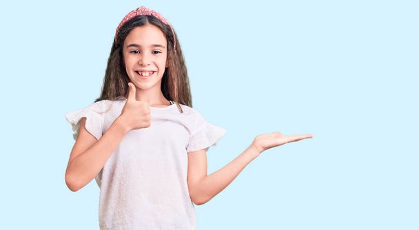 Cute hispanic child girl wearing casual white tshirt showing palm hand and doing ok gesture with thumbs up, smiling happy and cheerful  - Foto, afbeelding