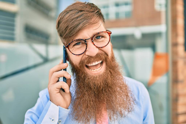 Giovane uomo d'affari rossa sorridente felice e parlando sullo smartphone in piedi alla città. - Foto, immagini