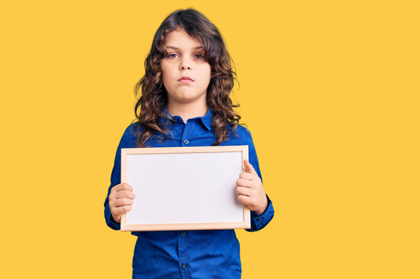 Cute hispanic child with long hair holding empty white chalkboard thinking attitude and sober expression looking self confident  - Foto, afbeelding