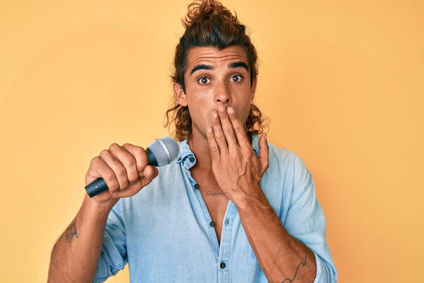 Young hispanic man singing song using microphone covering mouth with hand, shocked and afraid for mistake. surprised expression  - Foto, afbeelding