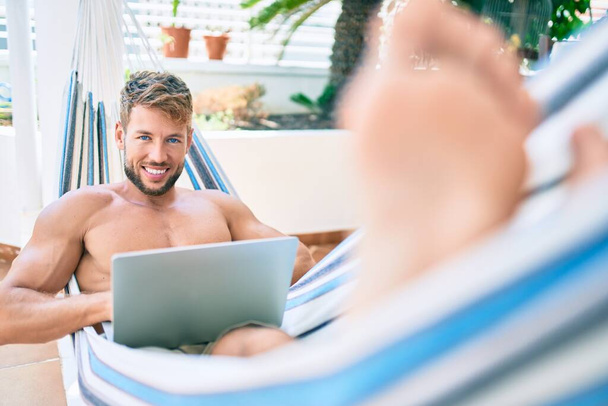 Jóképű fehér férfi mosolyog boldog pihenés egy függőágyon a teraszon számítógép laptop - Fotó, kép