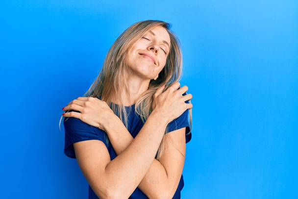 Mooie blanke vrouw draagt casual blauw t-shirt knuffelend zichzelf gelukkig en positief, glimlachend zelfverzekerd. zelfliefde en zelfzorg  - Foto, afbeelding