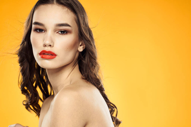 Beautiful brunette naked shoulders bright makeup red lips hairstyle close-up yellow background - Фото, изображение