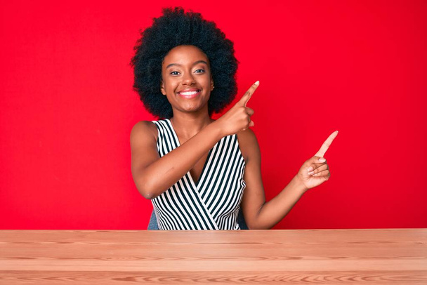 Genç Afro-Amerikalı kadın günlük kıyafetler giyiyor gülümsüyor ve kameraya bakıyor iki eliyle ve parmaklarıyla kenarı işaret ediyor..  - Fotoğraf, Görsel