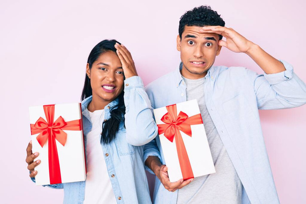 Beautiful latin young couple holding gift for anniversary celebration stressed and frustrated with hand on head, surprised and angry face  - Foto, imagen
