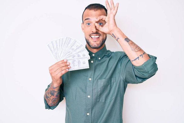 Young handsome man with tattoo holding dollars smiling happy doing ok sign with hand on eye looking through fingers  - Photo, image
