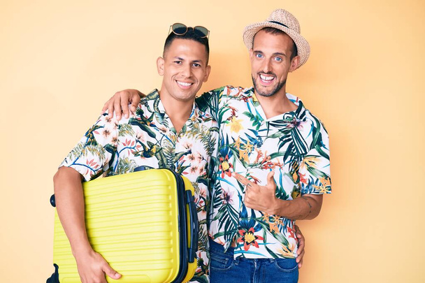 Νέοι ομοφυλόφιλοι δύο άνδρες κρατώντας βαλίτσα πηγαίνοντας καλοκαιρινές διακοπές χαμογελώντας ευτυχείς δείχνοντας με το χέρι και το δάχτυλο  - Φωτογραφία, εικόνα