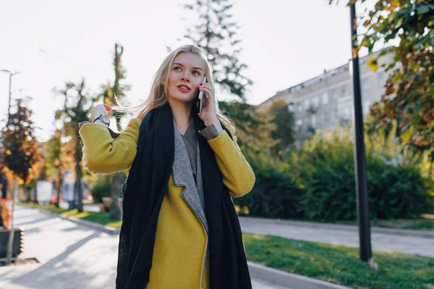 cute emotional attractive blonde woman in coat with smartphone walks the city street. communication during the walk, lifestyle, street in good weather. - Фото, изображение