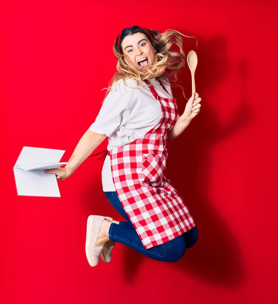 Young beautiful blonde cook woman wearing apron smiling happy. Jumping with smile on face holding recipe book and spoon over isolated red background - Foto, afbeelding