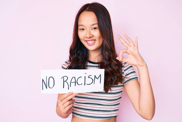 Νεαρή όμορφη Κινέζα κοπέλα που δεν κρατά πανό ρατσισμού κάνει ok υπογράψει με τα δάχτυλα, χαμογελώντας φιλικό gesturing εξαιρετικό σύμβολο  - Φωτογραφία, εικόνα