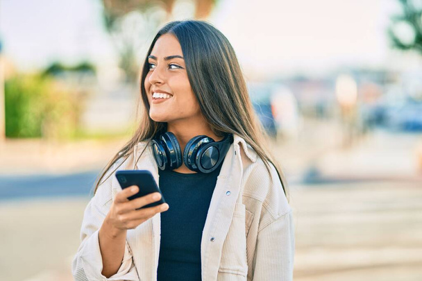 Joven chica hispana sonriendo feliz usando smartphone y auriculares en la ciudad. - Foto, imagen