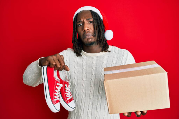 Afrikanischer Mann mit Zöpfen, Weihnachtsmütze und Schuhen als Geschenk, ahnungslos und verwirrt. Zweifelhaftes Konzept.  - Foto, Bild