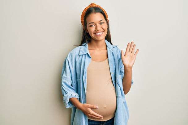 Beautiful hispanic woman expecting a baby, touching pregnant belly waiving saying hello happy and smiling, friendly welcome gesture  - Foto, Imagem