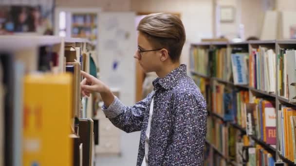 Un ragazzo con gli occhiali sceglie un libro in una biblioteca - Filmati, video
