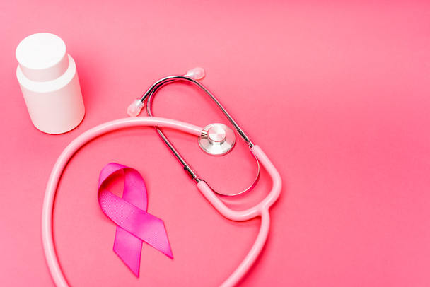 Stethoscope near pink ribbon of breast cancer awareness and jar with pills on pink background - Photo, Image