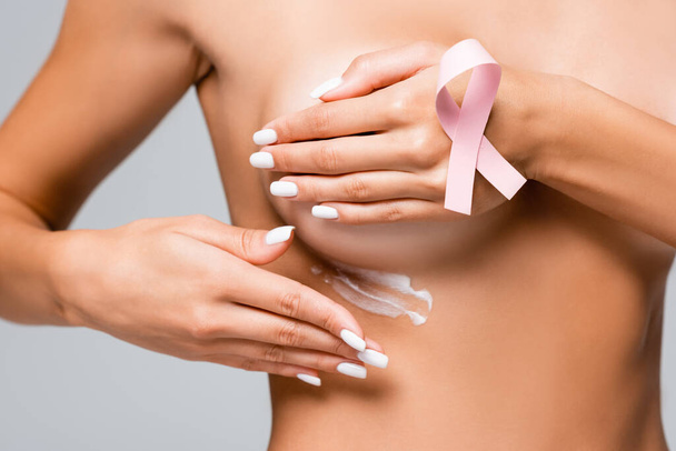 Vista recortada de mujer desnuda con cinta de conciencia de cáncer de mama aplicando crema cosmética aislada en gris  - Foto, imagen