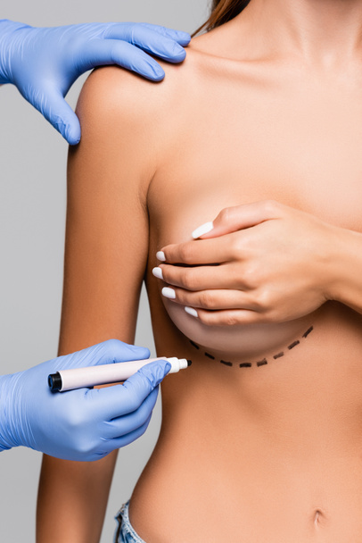 Άποψη του πλαστικού χειρουργού που κρατά το δείκτη κοντά στο στήθος της νεαρής γυναίκας που απομονώνεται στο γκρι  - Φωτογραφία, εικόνα