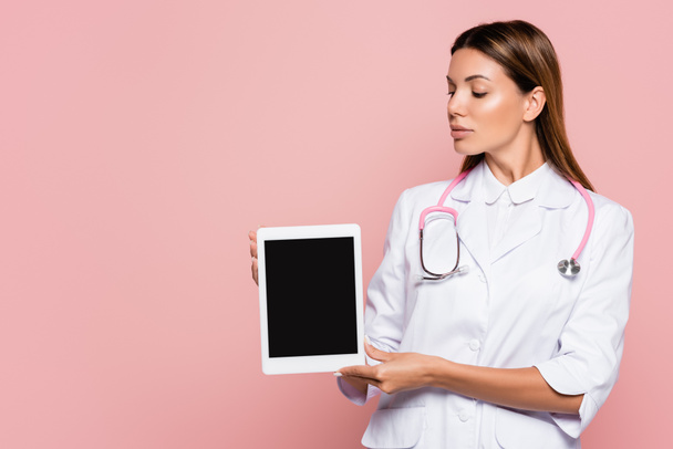 Doctor en bata blanca y estetoscopio sosteniendo tableta digital con pantalla en blanco aislado en rosa - Foto, Imagen