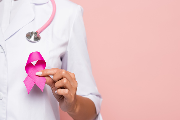 Vista recortada del médico sosteniendo el símbolo rosa de la conciencia del cáncer de mama aislado en rosa - Foto, imagen