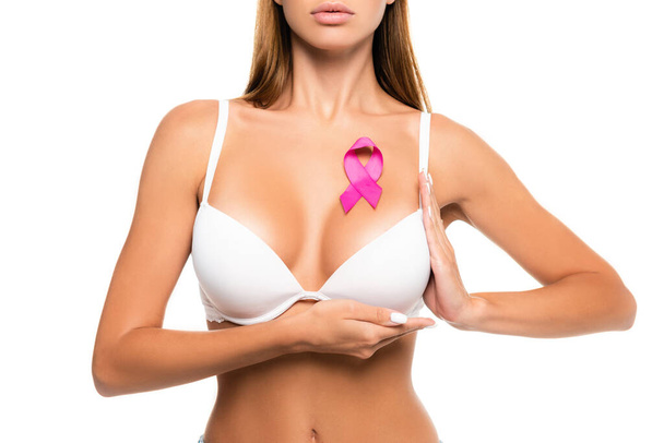 乳がんのピンクリボンを身につけながらブラジャーに触れる若い女性のクロップビューは白に隔離 - 写真・画像