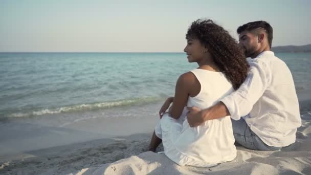 Romantische scène van een jong aantrekkelijk paar zit aan de kust te kijken naar de horizon bij zonsondergang - Mooie Spaanse vrouw leunt haar hoofd liefdevol op de schouder van haar knappe bebaarde vriendje - Video
