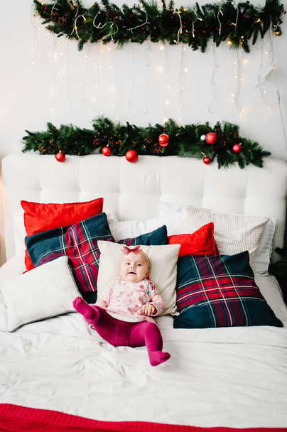 Baby girl on bed in bedroom near Christmas tree. Happy New Year and Merry Christmas. Christmas decorated interior. The concept of family holiday. - Фото, изображение