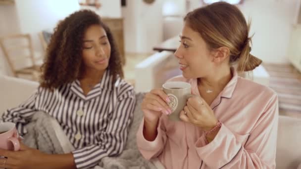 Indoor scene of two young beautiful women talking taking a cup of tea on the sofa under a blanket - Mixed race female couple  bonding each other at home sitting in  living room - Focus on right woman - Felvétel, videó