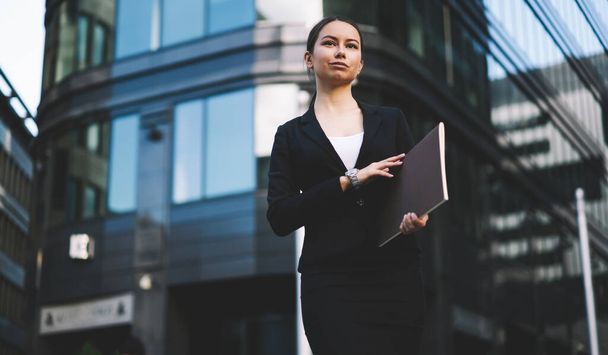 Low angle of elegant female in formal suit with folder with documents in hands and looking away near entrance of modern glass building - Foto, Bild