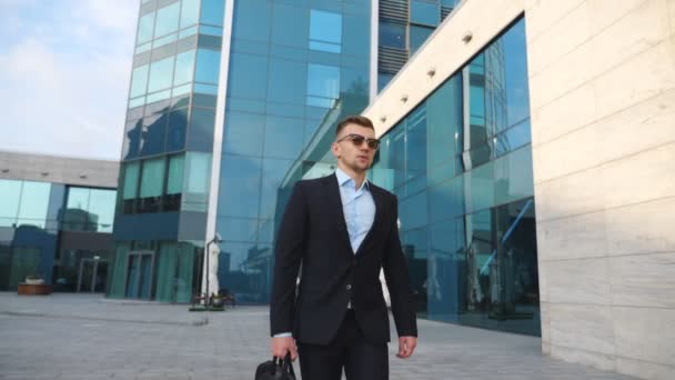 Giovane uomo d'affari in completo nero con valigetta che cammina lungo un moderno edificio per uffici. Fiducioso imprenditore maschio con borsa va a lavoro. Un bel ragazzo che va al lavoro. Rallentatore Close up - Filmati, video