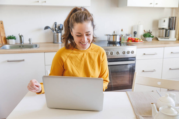 Oficina Móvil en casa. Mujer joven sentada en la cocina en casa trabajando con un ordenador portátil. Chica de estilo de vida estudiando o trabajando en interiores. Concepto de cuarentena empresarial independiente - Foto, imagen