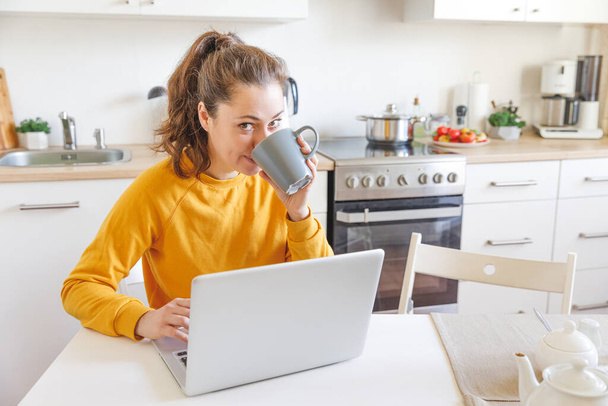 Κινητό γραφείο στο σπίτι. Νεαρή γυναίκα που κάθεται στην κουζίνα στο σπίτι και δουλεύει χρησιμοποιώντας φορητό υπολογιστή. Το κορίτσι του τρόπου ζωής σπουδάζει ή εργάζεται σε εσωτερικούς χώρους. Έννοια ελεύθερης επιχειρηματικής καραντίνας - Φωτογραφία, εικόνα