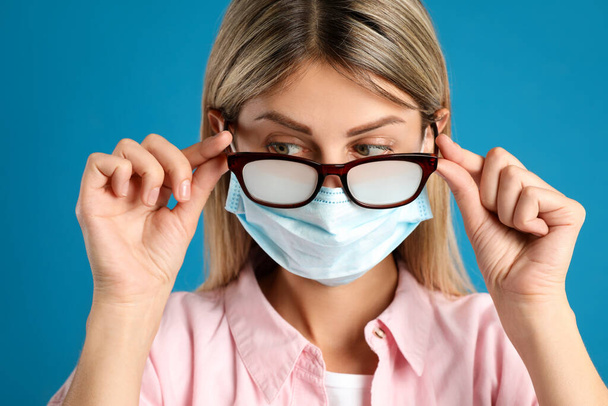 Woman with foggy glasses caused by wearing disposable mask on blue background. Protective measure during coronavirus pandemic - Φωτογραφία, εικόνα