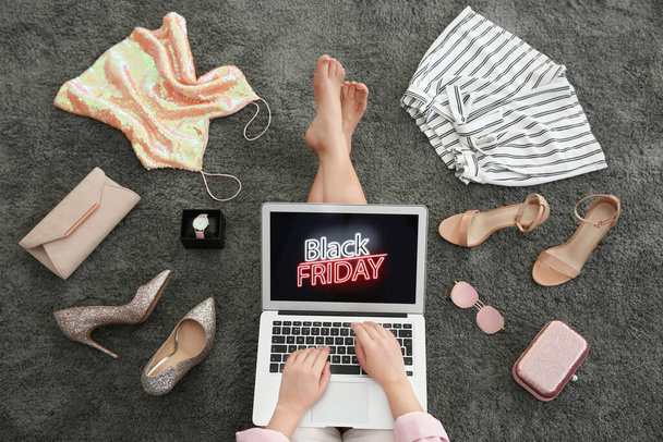 Woman surrounded by clothes and accessories using laptop with Black Friday announcement while sitting on grey carpet, top view - 写真・画像