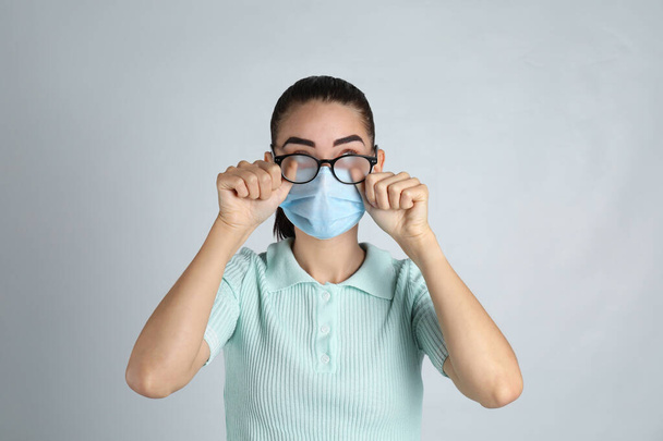 Woman wiping foggy glasses caused by wearing medical mask on light background - Foto, afbeelding