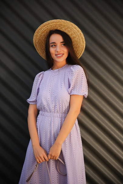 Beautiful young woman in stylish violet dress and straw hat near dark corrugated metal wall - Photo, image