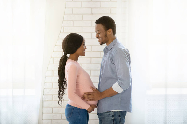 mooie zwart zwanger vrouw en haar man kijken naar elkaar met liefde thuis, zijaanzicht - Foto, afbeelding
