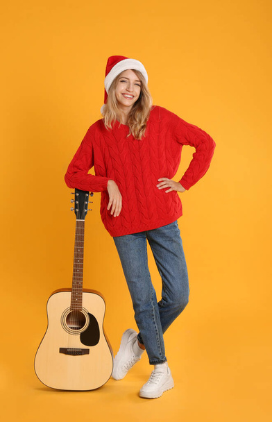 Young woman in Santa hat with acoustic guitar on yellow background. Christmas music - Zdjęcie, obraz