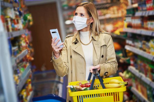 Dospělá žena v lékařské masce pomocí smartphonu a nakupování potravin - Fotografie, Obrázek