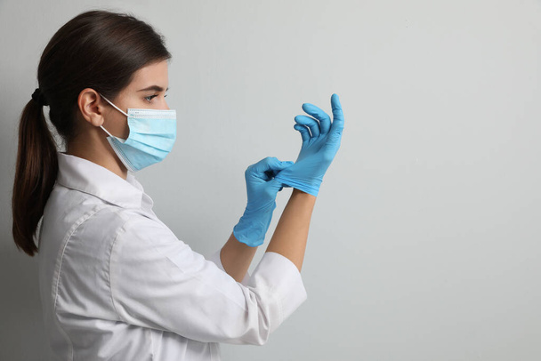 Doctor in protective mask putting on medical gloves against light grey background. Space for text - Zdjęcie, obraz