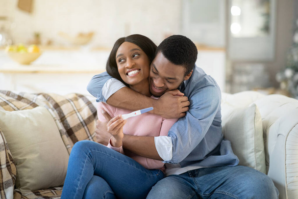 Liebender schwarzer Mann umarmt seine Frau zu Hause mit positivem Schwangerschaftstest - Foto, Bild