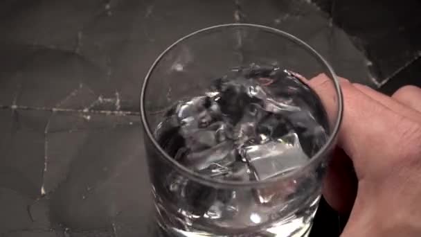 Kroužit vodou a ledem ve sklenici vody nebo alkoholu. Ledové kostky víří na černém vrásčitém vinobraní pozadí. Koncept starých nápojů. Zpomal. Zavřít - Záběry, video