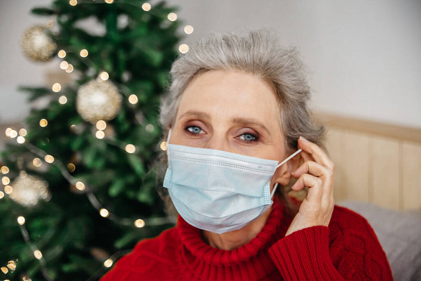Close up portrait of senior woman putting on mask at home christmas tree on background. Christmas at covid-19 concept. - Photo, Image