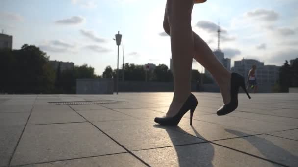 Slanke vrouwelijke benen in zwarte schoenen op hoge hakken lopen op het stadsplein bij zonsondergang. Voeten van jonge zakenvrouw in schoeisel op hoge hakken in stedelijke straat. Low angle view Slow motion Close up - Video