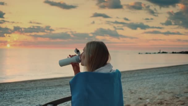 Fiatal nő iszik termál bögre és ül a strandon napkelte előtt - Felvétel, videó