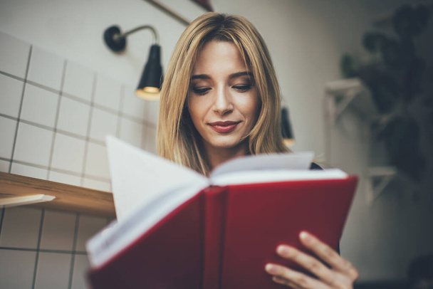 Attractive Caucasian female student enjoying leisure time for reading literature book with interesting plot, young hipster girl holding education textbook checking noted information for analyzing - Photo, image